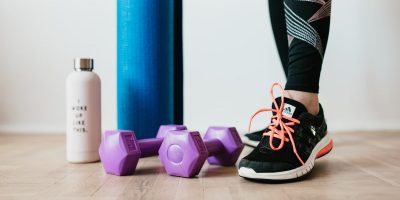 Crop sportswoman wearing sneakers and black leggings standing with sport mat on wooden floor near dumbbells and water bottle before exercising indoors